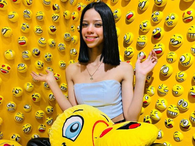 LiaBlom with straight black hair, smiling, wearing a strapless white top, hands up, in front of a yellow wall filled with emoji faces, holding a yellow emoji pillow.
