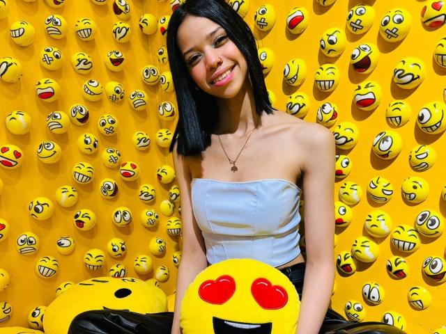 LiaBlom has small tits, black hair, smiling, wearing a white strapless top, holding a yellow emoji with red heart eyes, against a yellow wall of smiling emojis.