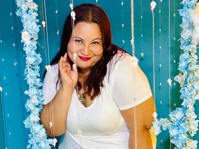 AliceShae with red hair, white shirt, smiling, holding a string of blue flowers, showing her tits and a tattoo on her chest, in front of a blue wall.