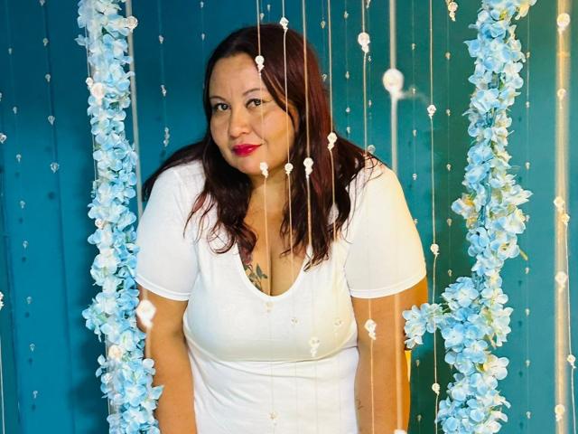 AliceShae, a chubby woman with brown hair, white shirt, and red lipstick, stands between blue floral and bead hangings, showing her small tits and tattoo on her chest.