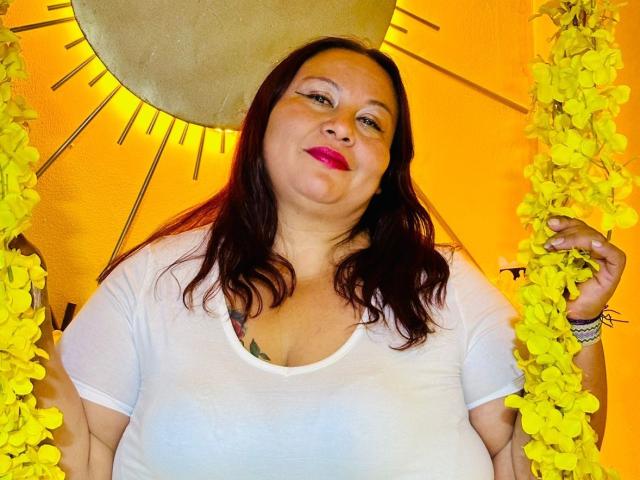 AliceShae has red lipstick, dark brown wavy hair, and a white t-shirt showing her large tits. She holds yellow flowers, smiling slightly, with a sun-like object behind.
