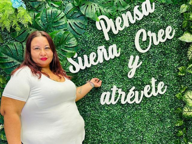 AliceShae with her big ass, full tits, and curvy body, wearing a white shirt, standing in front of a green leafy wall, smiling with red lipstick.