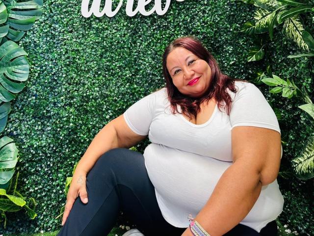 AliceShae has big tits, a fat belly, and a curvy ass, wearing a white tee and black pants, smiling with red lipstick, sitting against a green wall.