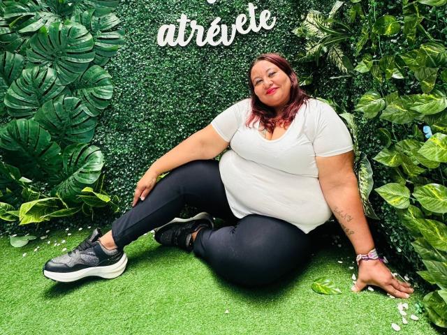 AliceShae, plus-sized with red hair, white tee, black pants, and black sneakers, sitting on green grass, her ass large, smiling, tattoo on right arm, leafy background.