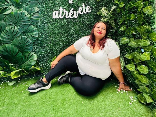 AliceShae, chubby with red hair, sitting on green grass, wearing a white tee, black pants, and black sneakers, against a lush green wall, smiling with red lipstick.