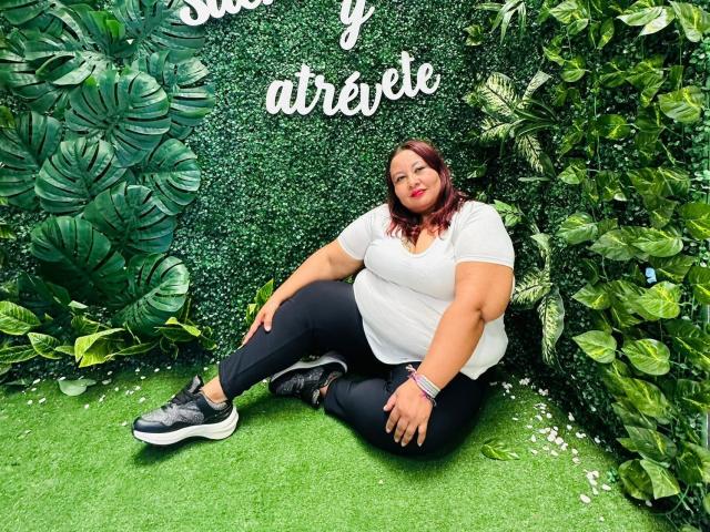AliceShae, plus-sized with large tits and ass, sits on green grass wearing a white tee and black pants, with dark sneakers, in front of a leafy wall.