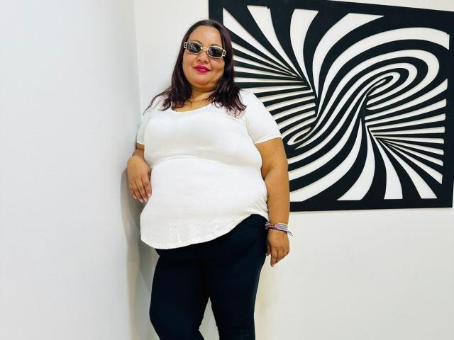 AliceShae with dark brown hair, wearing sunglasses, a white short-sleeve shirt, black pants, and pink lipstick, standing against a white wall with a black and white spiral pattern.