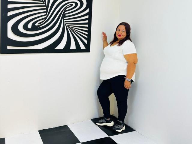 AliceShae stands in a white-walled room, leaning against the wall. She's curvy with a belly, wearing a white t-shirt, black pants, and black sneakers. Zebra art on wall.