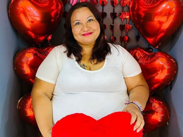 AliceShae, a chubby woman with dark brown hair, white shirt, and red heart pillow, surrounded by shiny red heart balloons, smiling with red lipstick.