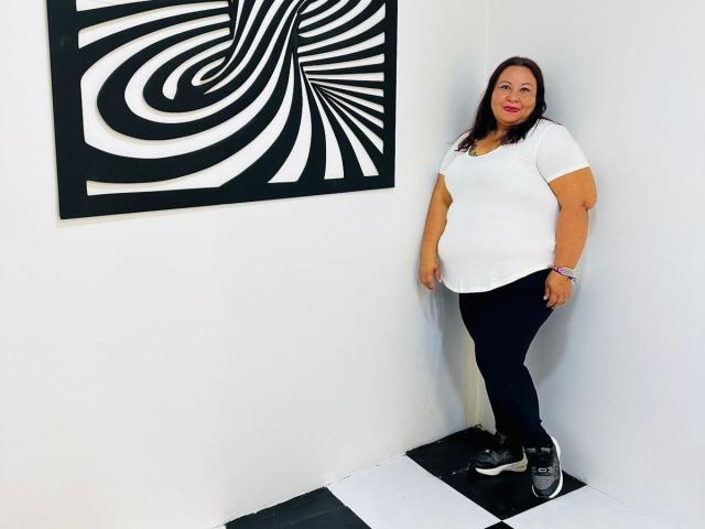 AliceShae has big tits, a curvy ass, and long dark hair, wearing a white shirt, black pants, and sneakers, standing in a corner with a black-and-white spiral painting.