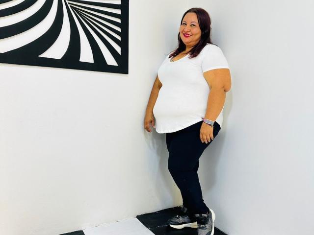 AliceShae, a chubby woman with red lipstick, wearing a white tee and black pants, stands in a white-walled room with black-and-white wall art. She's smiling.
