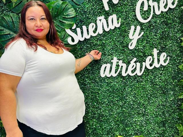 AliceShae has large tits, a chubby ass, and a white shirt, standing against a green wall, with red lipstick and brown hair, smiling.