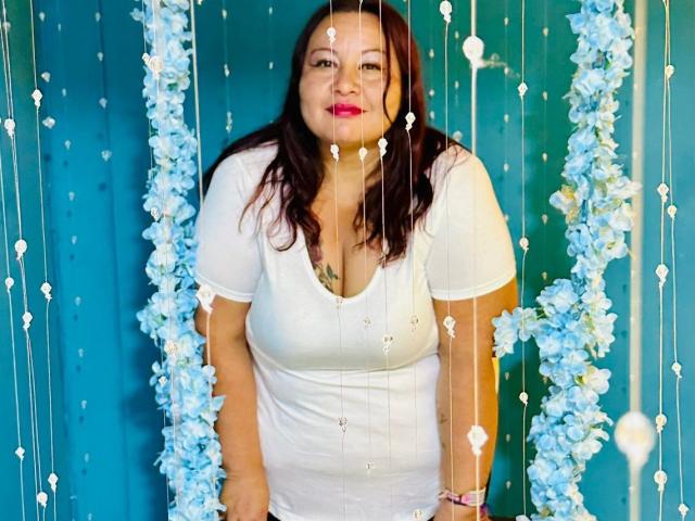 AliceShae has red hair, white dress, medium tits, standing with blue floral garlands and white beads hanging in front, smiling with pink lipstick, against a teal background.