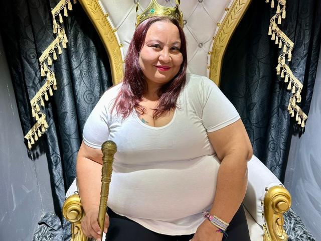 AliceShae, chubby with red hair, white tee, and black pants, sits on a gold throne, holding a gold staff, wearing a gold crown, smiling at the camera.