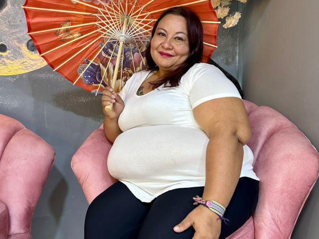 AliceShae, chubby with big tits, wearing a white shirt and black pants, holding an orange paper fan, sitting in a pink chair, smiling with red lipstick.