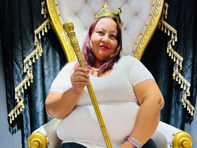 AliceShae, chubby with pinkish-brown hair, sits on a golden throne holding a golden staff, wearing a white tee, smiling with red lipstick, and has a bracelet on her wrist.