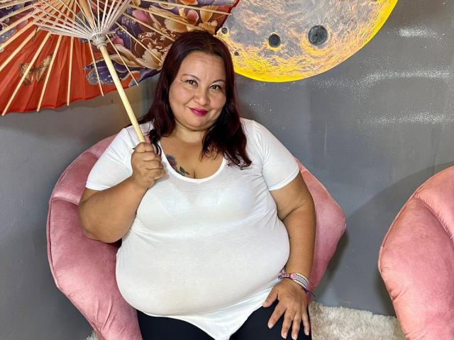AliceShae with red hair, white shirt, black pants, holding red paper umbrella, sitting on pink chair, smiling, chubby, tattoo on chest, pink wristband, gray wall, pink chair.
