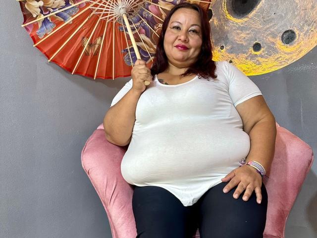 AliceShae is a chubby woman with red hair, wearing a white shirt and black pants, sitting on a pink chair, holding a red and gold fan, smiling.