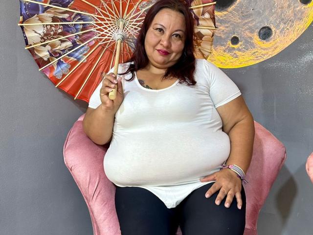 AliceShae, a chubby woman with red hair, holds a colorful fan, wearing a white shirt and black pants, sitting on a pink chair, smiling.