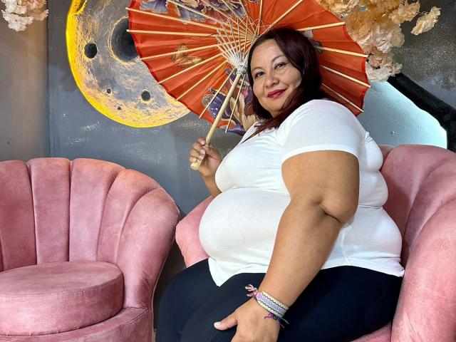 AliceShae with red hair, white shirt, black pants, holding orange fan, sitting on pink chair, smiling, curvy, large tits, chubby, pink bracelet, black armchair, floral wall decor.