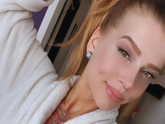 PixiFleur-hot has light skin, blonde hair in a ponytail, a nose ring, and a small smile. She's wearing a white robe with a pink top underneath.