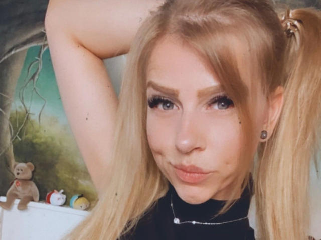 PixiFleur-hot with puffy lips, blonde twintails, and black ear gauges, wearing a black top, arms raised, showing a cute stuffed teddy bear in the background.