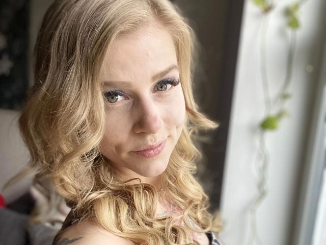 PixiFleur-hot has blonde curls, green eyes, and a subtle nose ring, smiling with her tits slightly visible, standing near a window with a string of leaves.