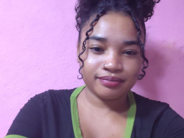 HellaTinot has curly black hair in a top knot, green V-neck shirt, black top, nose ring, and a subtle smile. Pink wall in background. Shiny, smooth skin.