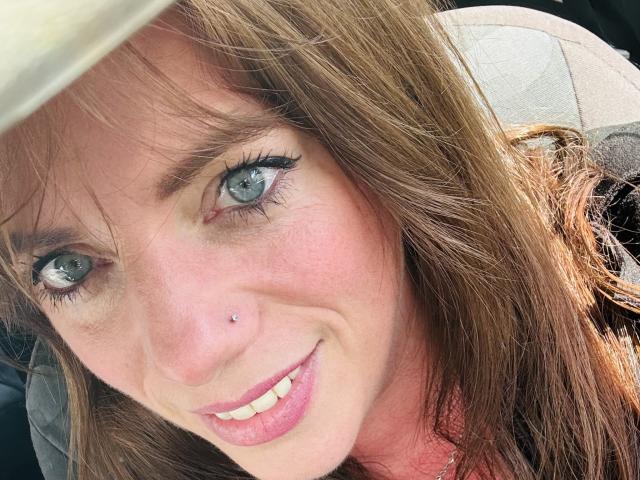 SerenaFr with blue eyes, brown hair, and a nose piercing, smiling at the camera, wearing a beige hat, and a gray jacket, showing her white teeth and pink lips.