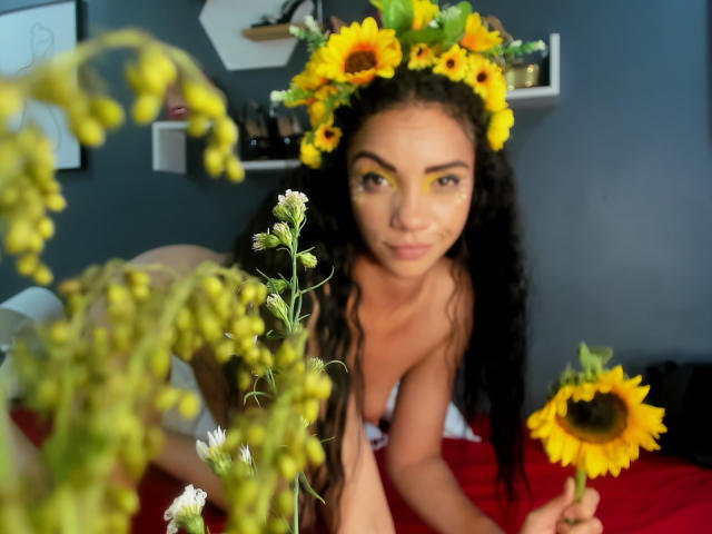 AndromedaZareth with sunflower headband, yellow eye makeup, holding sunflower, lying on red bed, surrounded by green and white flowers, curly black hair, looking at camera, nude.