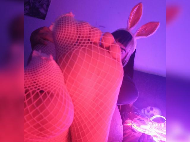 Lxlitx with pink fishnet stockings on her feet, bunny ears, and purple lighting, showing her soles up, in a sexy, playful pose, with her face partially visible.
