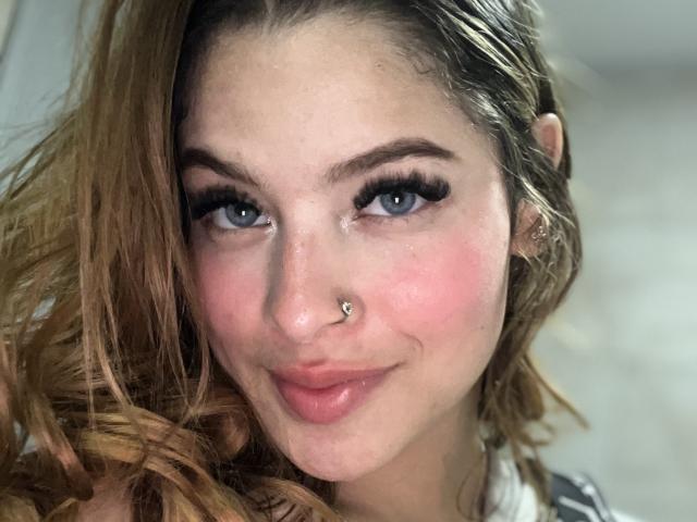 ShantalSummer has blue eyes, a nose ring, and long wavy brown hair. She's smiling with pink cheeks and full lips, wearing black eyeliner and mascara.