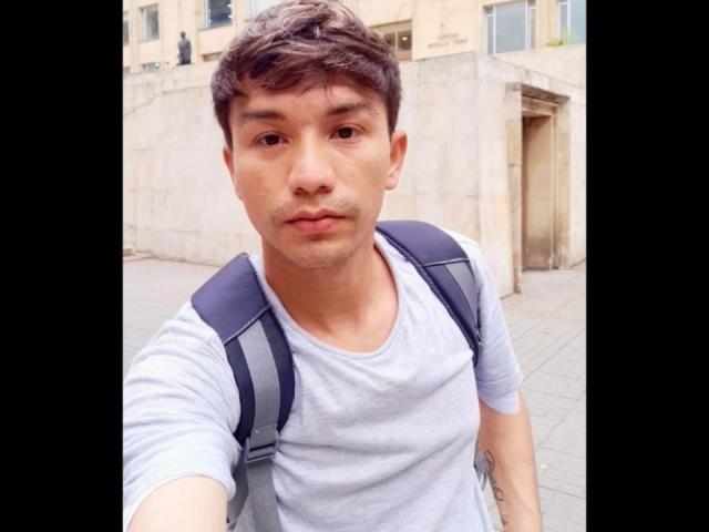 FlacoVergon69 with short brown hair, light skin, wearing a gray shirt, black backpack, and a neutral expression, taking a selfie. Tattoo on right arm visible.