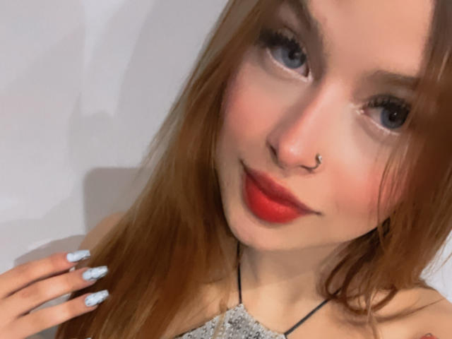 ShantalSummer with red lipstick, nose ring, and blue eyes, smiling slightly, showing off long red hair and white nails with blue tips, wearing a sparkly top.