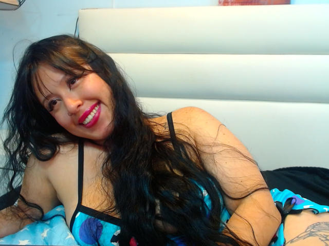 AmyHarriis with long black hair, smiling, wearing a blue and black lingerie set, showing her tits, lying on a bed with a blue star-patterned blanket.
