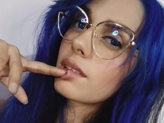 Anais Hilton mit blauen Haaren, die eine Brille trägt, sich an ihren Finger beißt, ein leichtes Lächeln zeigt und verführerisch in die Kamera schaut. Ihre Brüste sind teilweise sichtbar..