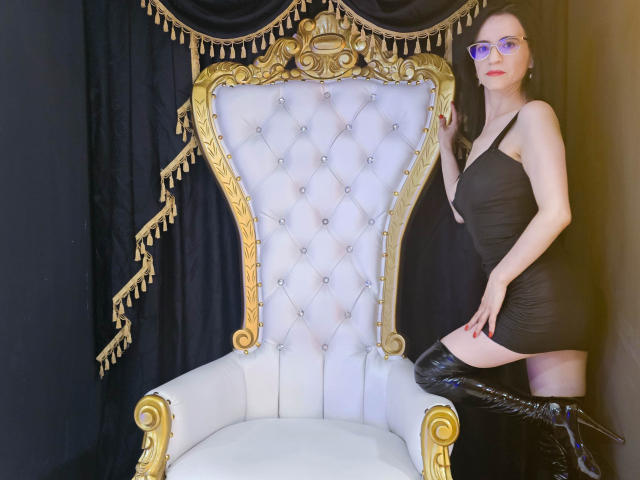AlanaMoneth in a black dress, purple glasses, and black thigh-high boots, kneeling on a gold and white tufted throne, hand on throne, looking at camera.