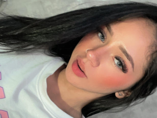 KloeThompson has black hair, blue eyes, pink blush, and slightly parted pink lips. She wears a white shirt with pink details. Her nose is small and she looks at the camera.