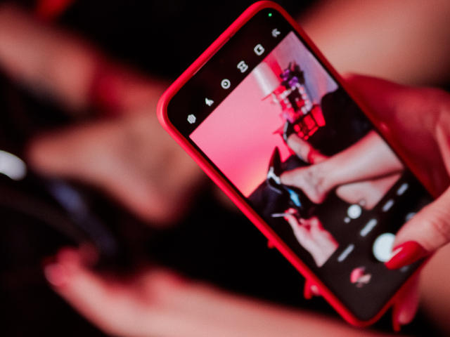 NicoleDomme's hand holding a red phone, showing a selfie of her feet in black high heels with red nails, blurred background, focused on phone screen, sexy.