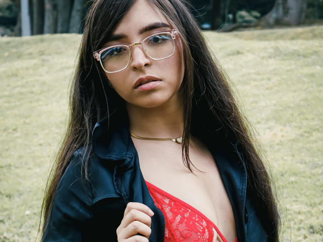 EvaMcAllister with long brown hair, wearing glasses, black jacket, and red lace bra, exposing her tits, standing in a grassy area, looking serious.