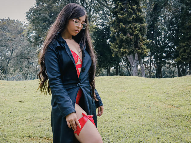 EvaMcAllister with long brown hair, glasses, black leather coat, red strappy lingerie, and garter, standing in grassy field, looking down, sexy, attractive, exposed breasts, visible pussy strap.