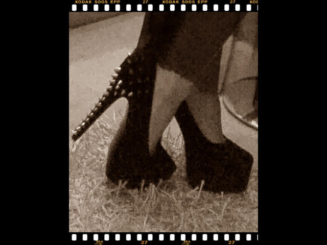 Amybabyporn-hot's ass in dark, spiked high heels, showing off her thick, black-clad legs and a studded ankle strap, standing on grassy ground in a sepia-toned shot.
