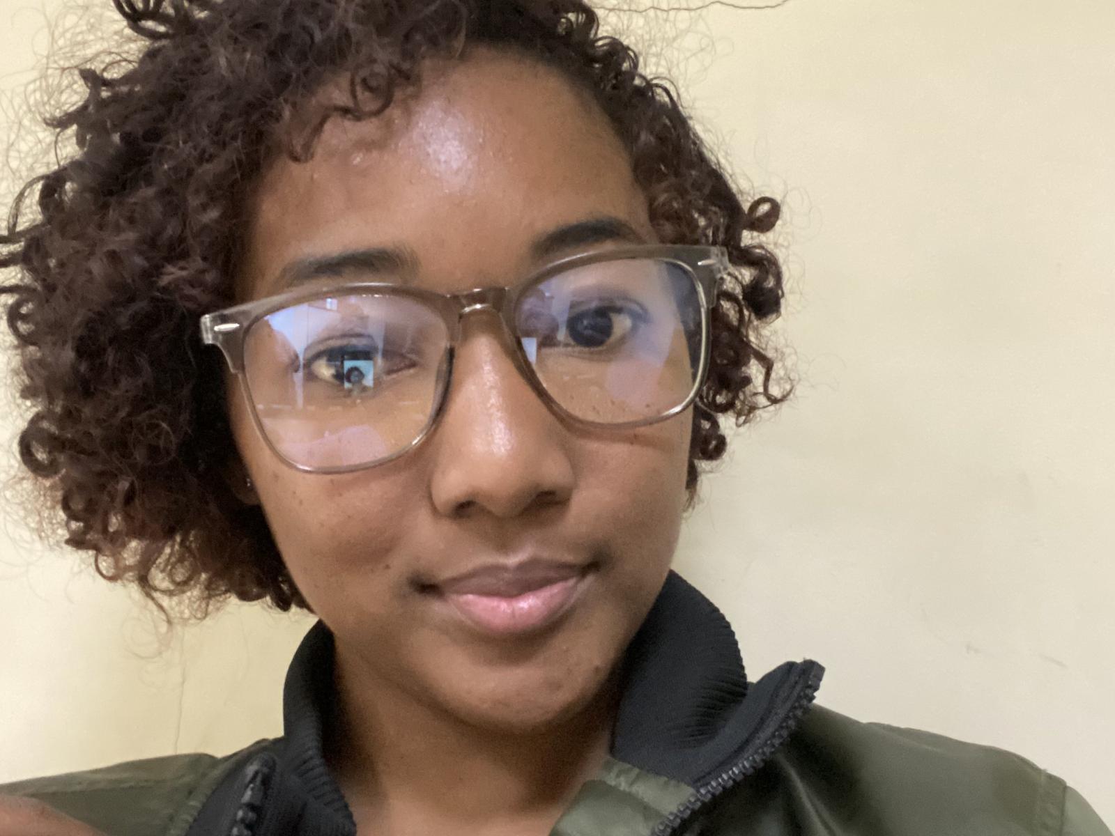 CassieJolie with curly brown hair, wearing clear glasses and a green jacket, has a slight smile and shiny brown skin. Her small ass is partially visible.
