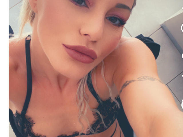 Alisssa has blonde hair, purple eyelids, and dark lipstick. She's wearing a black lace bra, showing her tits, with a tattoo on her right arm.