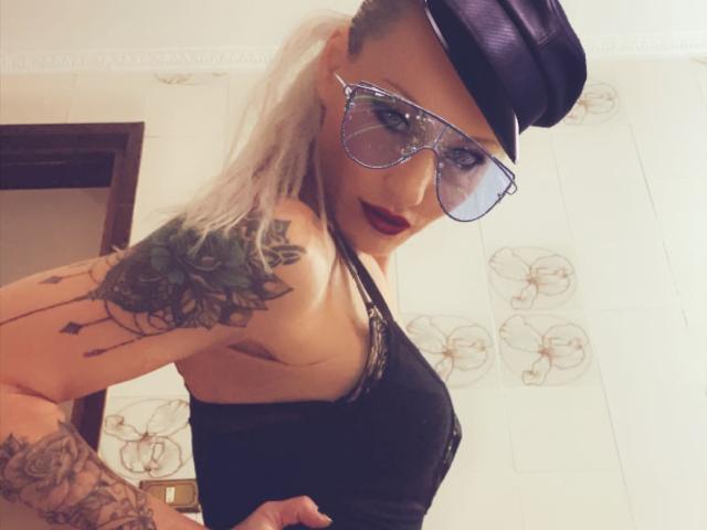 Alisssa with tattoos, glasses, and a black hat, wearing a black top, showing her tits and a side view of her ass. Her blonde hair is in a ponytail.
