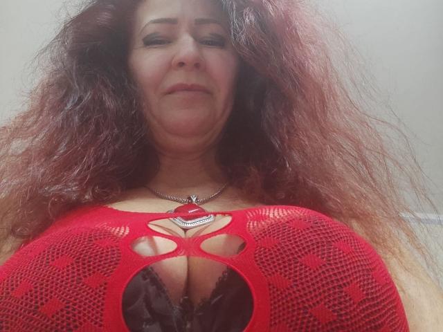 MilayaDFlow, avec ses cheveux roux et ondulés, portant un haut rouge en résille qui expose ses seins volumineux et foncés, souriant légèrement, debout devant un fond uni..