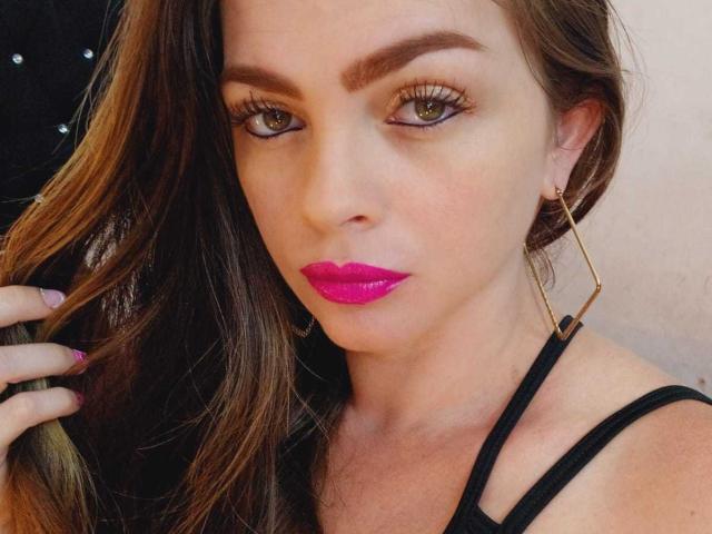 KittyBonx has bright pink lipstick, green eyes, and brown hair, wearing gold earrings and a black strap top, with a serious expression and hand in her hair.