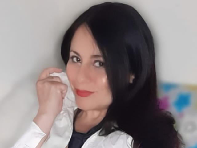 EkaterinaSex with black hair, white lab coat, red lipstick, and black eyes, smiling slightly, holding her chin with her right hand, wearing a black shirt.