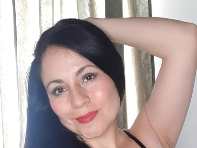 EkaterinaSex with black hair, fair skin, red lipstick, and brown eyes, raising her right arm, showing her tits, smiling slightly, against a light-colored curtain.