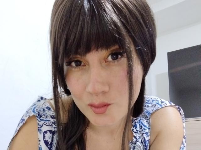 AngelCams with dark brown hair and bangs, pouty pink lips, and blue patterned top, looking at the camera with slightly raised eyebrows. She has big, dark eyes.